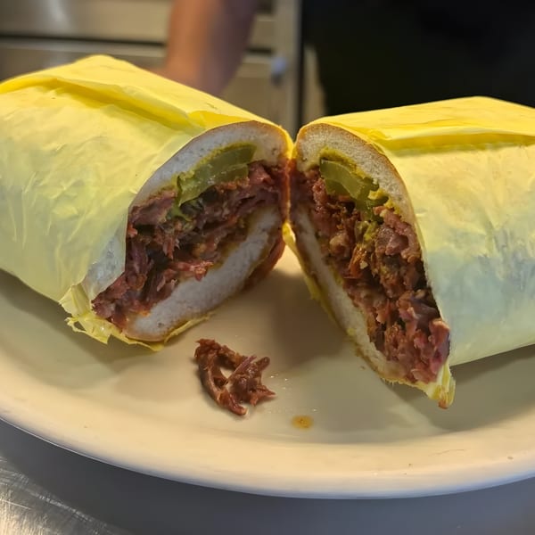 A Jim's Burgers & Eggs pastrami sandwich, cut in half, wrapped in yellow paper on a white plate.