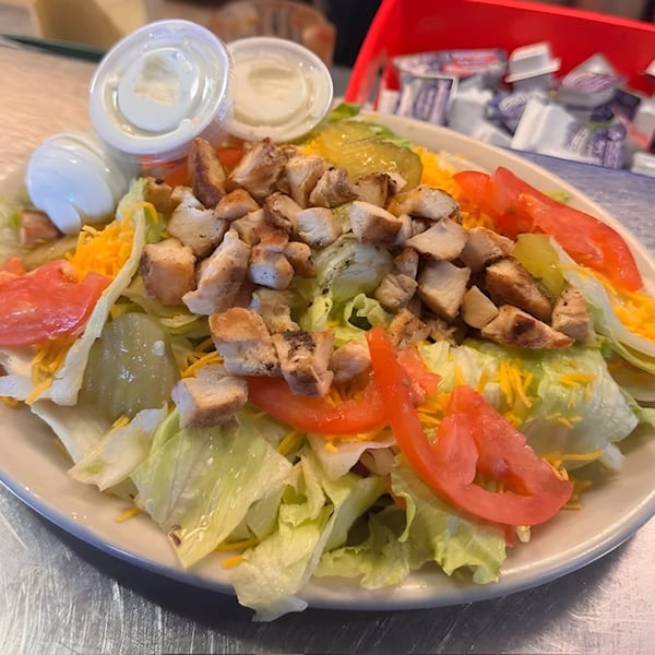 A grilled chicken salad with lettuce, tomatoes, cheese, pickles, and dressing cups on a white plate.