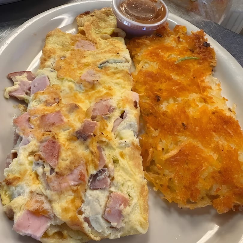 A ham and cheese omelet with a side of crispy hash browns and a small cup of sauce.