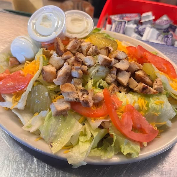 A grilled chicken salad with lettuce, tomatoes, shredded cheese, and pickles, with two dressing cups.